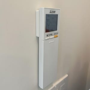 Może przedstawiać: Biały pilot do klimatyzacji Mitsubishi Electric zamontowany na ścianie. Pilot posiada cyfrowy wyświetlacz pokazujący temperaturę oraz przyciski sterowania z napisami "TEMP" i "OFF/ON".