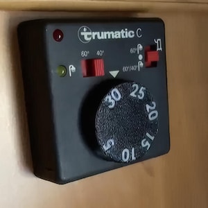 Könnte beinhalten: Ein schwarzes Trumatic C Bedienfeld mit rundem Drehknopf und roten und grünen Kontrollleuchten. Das Zifferblatt hat Zahlen von 5 bis 30. Es gibt rote Schalter und das Trumatic-Logo.