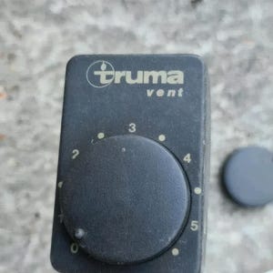Peut inclure: Un panneau de commande de ventilation Truma noir avec un cadran circulaire et les chiffres de 0 à 5. Le mot "Truma" est imprimé au-dessus de "vent". Un petit capuchon noir rond se trouve à droite du panneau.