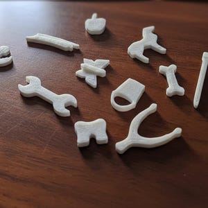 Pode incluir: Uma coleção de pequenos objetos brancos impressos em 3D espalhados sobre uma superfície de madeira. Os objetos incluem uma chave inglesa, um cão, um osso, uma dedeira, um amuleto e outras formas. Os objetos são todos de cor neutra.