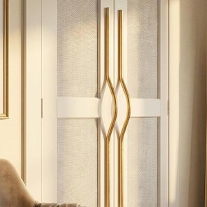 May include: A pair of long, gold-toned metal door handles with a curved, wave-like center design mounted vertically on white double doors featuring textured, light-colored woven panels. The handles have a smooth, polished finish.