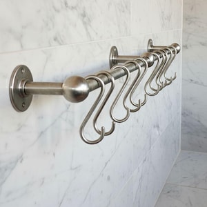 May include: A brushed nickel metal pot rack with eight S-hooks, mounted on a white marble tile wall. The rack has a round ball on the end and is attached to the wall with circular mounting plates.