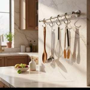 Puede incluir: Un estante de utensilios de cocina de acero inoxidable con herramientas colgantes. Las herramientas incluyen una cuchara de madera, un cucharón, un batidor y pinzas. El estante está montado en una pared de mármol.