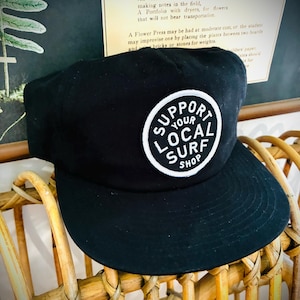 May include: Black baseball cap with a flat brim. The cap features a white circular patch with the text "SUPPORT YOUR LOCAL SURF SHOP" in a black, bold font. The hat is sitting on a wicker chair.