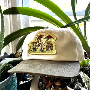 May include: A beige baseball cap with a green brim. The cap features a patch with a mushroom design in red, brown, and yellow. The hat is made of a soft material and is suitable for casual wear.