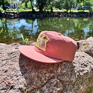 May include: A dusty rose-colored baseball cap with a curved brim, resting on a rock. The cap features a patch with a mushroom design in shades of red, yellow, and brown. The background includes a body of water and trees.
