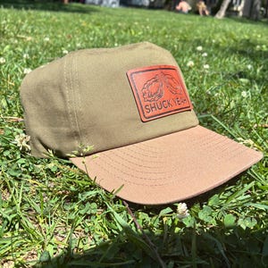 May include: A khaki baseball cap with a brown bill and a brown leather patch. The patch features an oyster design and the text "SHUCK YEAH". The cap is resting on green grass.