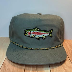 Op de afbeelding: Een groene pet met een kleurrijke geborduurde vispatch. De vis heeft rode, groene en gele details. De rand heeft een touwtrim met een camouflagepatroon.