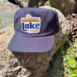 May include: Navy blue baseball cap with a curved brim. The cap features a patch with the words "Lake Mode" in blue and gold, with a graphic of a can. The cap is sitting on a rock.