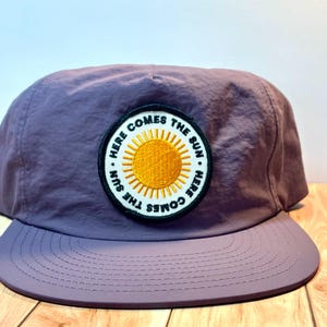Può includere: Un cappellino da baseball viola con visiera piatta. Il cappellino presenta una toppa circolare con la scritta "HERE COMES THE SUN" attorno a una grafica solare gialla. Il cappello è realizzato in un materiale leggero.