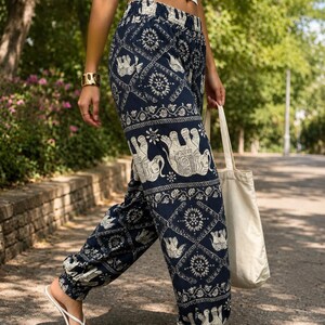 May include: Navy blue loose-fitting trousers featuring a repeating white elephant and floral mandala pattern. The trousers have an elasticated waistband and elasticated cuffs at the ankles, paired with a plain white crop top and a simple cream tote bag.
