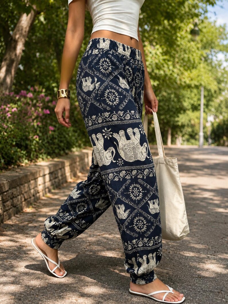 May include: Navy blue loose-fitting trousers featuring a repeating white elephant and floral mandala pattern. The trousers have an elasticated waistband and elasticated cuffs at the ankles, paired with a plain white crop top and a simple cream tote bag.