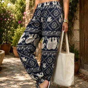 May include: Navy blue loose-fitting trousers featuring a white elephant and geometric pattern, an elastic waistband, and elasticated ankle cuffs. A plain white crop top and a simple cream-coloured canvas tote bag accompany the patterned trousers.