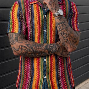 May include: A short-sleeved button-down shirt featuring a vibrant vertical striped pattern in shades of yellow, red, green, blue, and purple. The shirt has a classic collar, a front button placket with white buttons, and a textured fabric finish.