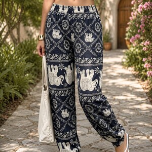 May include: High-waisted loose-fitting trousers in a dark navy blue fabric featuring a repeating white pattern of elephants and geometric floral motifs. The trousers have an elasticated waistband and elasticated cuffs at the ankles, paired with a white tote bag.