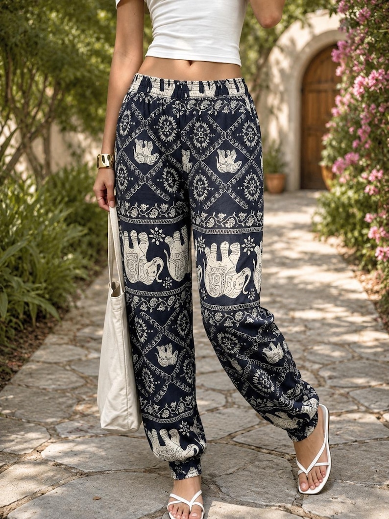 May include: High-waisted loose-fitting trousers in a dark navy blue fabric featuring a repeating white pattern of elephants and geometric floral motifs. The trousers have an elasticated waistband and elasticated cuffs at the ankles, paired with a white tote bag.