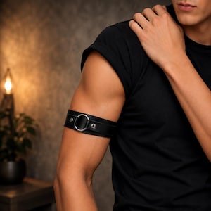 May include: A black leather armband featuring a central silver-toned metal ring. The band is secured with two silver-toned metal rivets on either side of the ring, creating a smooth, circular design that wraps around the upper arm.