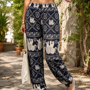 May include: High-waisted trousers featuring a dark blue base with a repeating white pattern of elephants and intricate geometric floral motifs. The trousers have an elasticated waistband and elasticated cuffs at the ankles, paired with a plain white crop top.