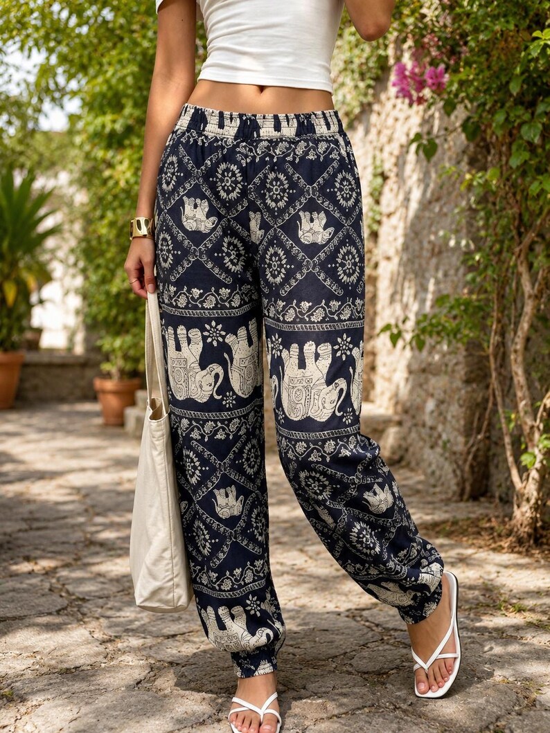 May include: High-waisted trousers featuring a dark blue base with a repeating white pattern of elephants and intricate geometric floral motifs. The trousers have an elasticated waistband and elasticated cuffs at the ankles, paired with a plain white crop top.