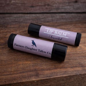 K&ouml;nnte beinhalten: Zwei Lippenbalsam-Tuben mit schwarzen Kappen und lavendelfarbenen Etiketten. Auf einem Etikett steht "LIP BALM Unscented". Auf dem anderen Etikett steht "Farmers Daughter Tallow Co." mit einer blauen Vogelgrafik. Die Tuben liegen auf einer Holzoberfl&auml;che.