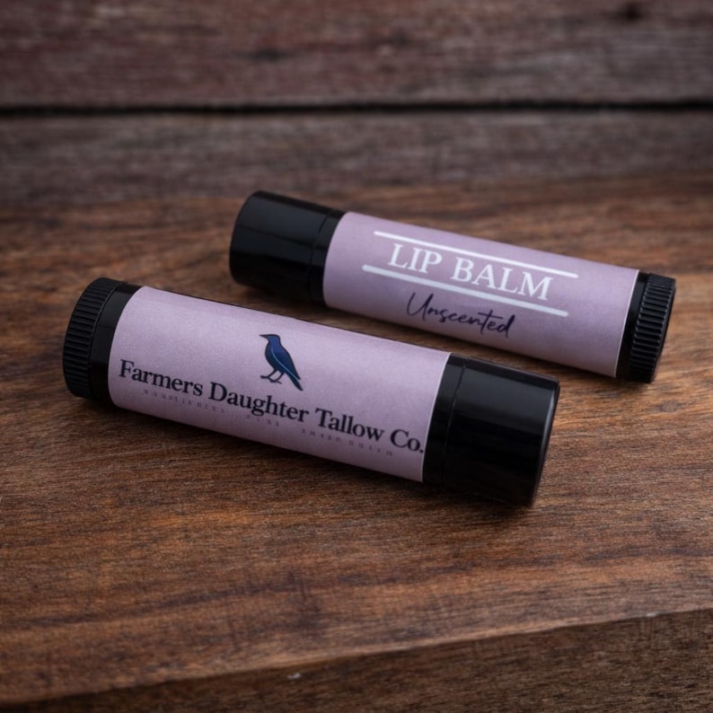K&ouml;nnte beinhalten: Zwei Lippenbalsam-Tuben mit schwarzen Kappen und lavendelfarbenen Etiketten. Auf einem Etikett steht "LIP BALM Unscented". Auf dem anderen Etikett steht "Farmers Daughter Tallow Co." mit einer blauen Vogelgrafik. Die Tuben liegen auf einer Holzoberfl&auml;che.