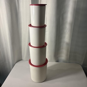 May include: A stack of four white cylindrical food storage containers with red lids. The containers are arranged vertically, with the largest at the bottom and the smallest at the top. The containers are made of a smooth, opaque material.