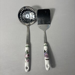 May include: A stainless steel slotted spoon and spatula set. The handles are white with floral designs in purple, pink, and green. The spoon has a round head with multiple holes. The spatula has a rectangular head.