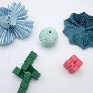 Könnte beinhalten: Eine Auswahl an farbenfrohen geometrischen Objekten. Ein hellblaues, blütenförmiges Objekt, ein teal-farbenes sternförmiges Objekt, ein grüner Ball, ein grüner, beweglicher Block und ein rosafarbener Würfel sind auf einer weißen Oberfläche angeordnet.