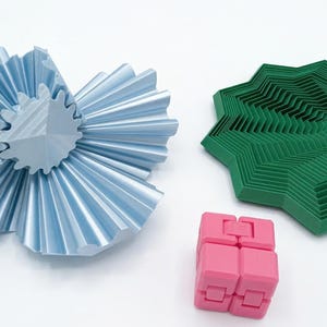 Pode incluir: Uma variedade de objetos geométricos impressos em 3D em várias cores. Uma estrutura azul clara em forma de flor com um centro em forma de engrenagem está ao lado de um objeto verde em forma de estrela. Um brinquedo fidget rosa em forma de cubo também está presente.