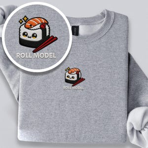 May include: A heather gray crewneck sweatshirt featuring an embroidered design of a smiling sushi roll with a salmon slice on top, red chopsticks, and two yellow stars. The text ROLL MODEL is embroidered in white capital letters below the sushi.