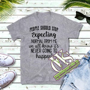 May include: A heather gray t-shirt with the text "PEOPLE SHOULD STOP Expecting NORMAL FROM ME we all know it's NEVER GOING TO happen!" is displayed. The shirt is paired with blue jeans, white sneakers, and sunglasses.