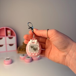 May include: A pink handheld digital pet game with a white bow and screen displaying two pixelated hearts. The game is attached to a silver clasp. Other pink accessories are in the background.