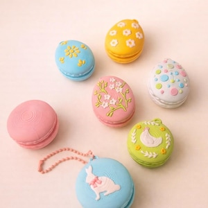 May include: An assortment of colorful, Easter-themed macarons. The macarons are decorated with floral and bunny designs, and egg shapes. Colors include blue, yellow, pink, green, and white. One macaron has a pink chain.