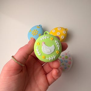 May include: A hand holds four colorful Easter egg ornaments. The ornaments are in shades of blue, yellow, green, and pink. The green ornament features a white chicken design. The other ornaments have floral patterns.