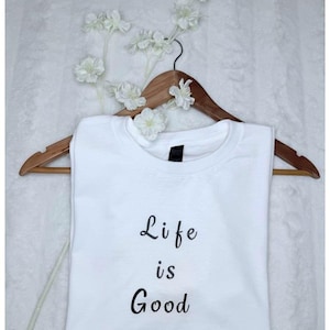 May include: A white t-shirt with the phrase "Life is Good" in black script. The shirt is displayed on a wooden hanger, adorned with white floral accents. The overall aesthetic is simple and clean.