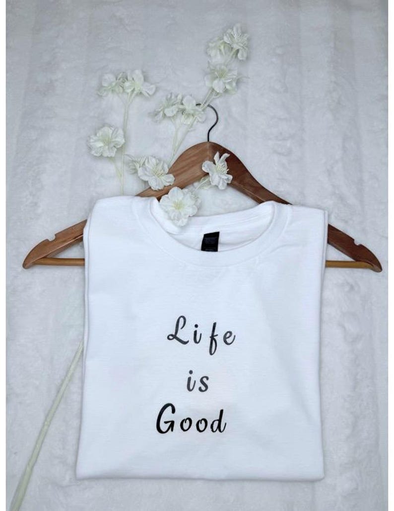 May include: A white t-shirt with the phrase "Life is Good" in black script. The shirt is displayed on a wooden hanger, adorned with white floral accents. The overall aesthetic is simple and clean.