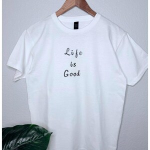 May include: A white t-shirt with the phrase "Life is Good" printed in black script. The shirt is hanging on a wooden hanger against a light gray wall. A green plant is visible in the lower left corner.