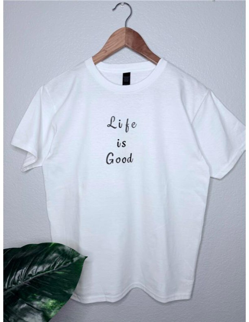 May include: A white t-shirt with the phrase "Life is Good" printed in black script. The shirt is hanging on a wooden hanger against a light gray wall. A green plant is visible in the lower left corner.