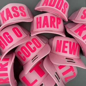 May include: A collection of pink trucker hats with various neon pink text, including "KASS", "HARP", "COCO", and "NEWT". The hats have a mesh back and a curved brim. The hats are stacked together.