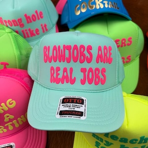 May include: A collection of trucker hats in various colors, including teal, pink, blue, and green. Each hat features bold, eye-catching text in contrasting colors. The hats have a mesh back and a foam front, with a snap closure.