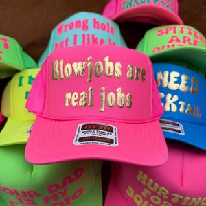 May include: A collection of trucker hats in various bright colors, including pink, green, and blue. Each hat features bold text in contrasting colors, with phrases like "Blowjobs are real jobs" and "Big Dick Inspector". The hats have a mesh back and a snap closure.