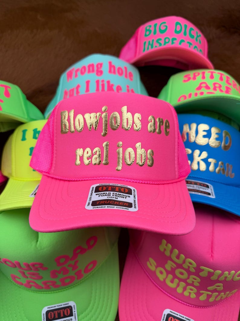 May include: A collection of trucker hats in various bright colors, including pink, green, and blue. Each hat features bold text in contrasting colors, with phrases like "Blowjobs are real jobs" and "Big Dick Inspector". The hats have a mesh back and a snap closure.