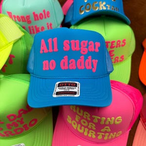 May include: A collection of trucker hats in various bright colors, including blue, pink, green, and yellow. Each hat features bold text in contrasting colors, with phrases like "All sugar no daddy" and "I need cocktail".