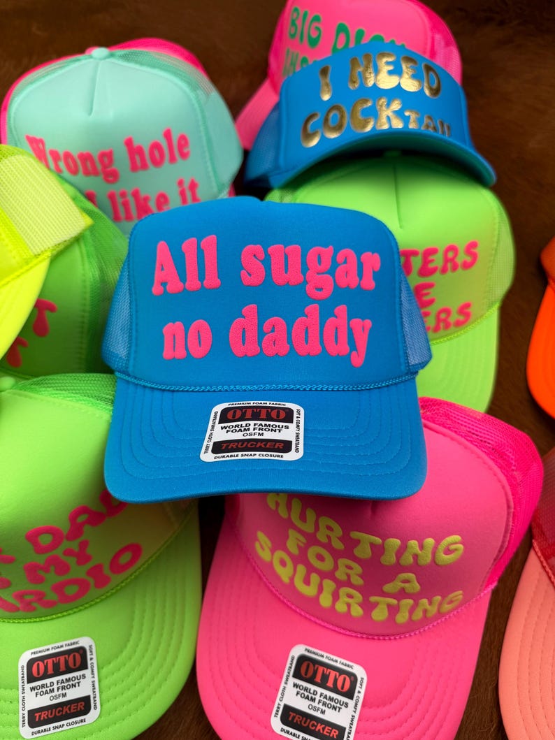 May include: A collection of trucker hats in various bright colors, including blue, pink, green, and yellow. Each hat features bold text in contrasting colors, with phrases like "All sugar no daddy" and "I need cocktail".
