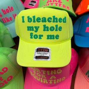 May include: A collection of trucker hats in various bright colors, including neon green, pink, and blue. Each hat features bold, humorous text in contrasting colors. The hats have a mesh back and a snap closure.