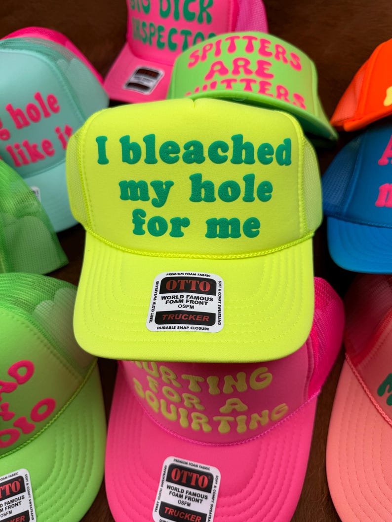 May include: A collection of trucker hats in various bright colors, including neon green, pink, and blue. Each hat features bold, humorous text in contrasting colors. The hats have a mesh back and a snap closure.