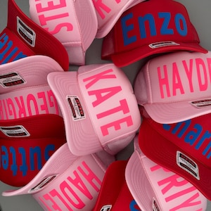 May include: A collection of trucker hats in various colors, including pink and red. Each hat features a name printed in a contrasting color across the front. The hats have a mesh back and a curved brim.
