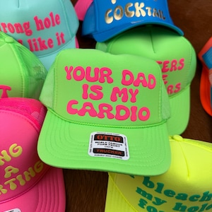 May include: A collection of trucker hats in various bright colors, including neon green, pink, and blue. Each hat features bold, humorous text in contrasting colors, such as "Your Dad Is My Cardio" and "I Need Cocktail".