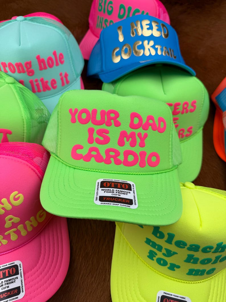 May include: A collection of trucker hats in various bright colors, including neon green, pink, and blue. Each hat features bold, humorous text in contrasting colors, such as "Your Dad Is My Cardio" and "I Need Cocktail".