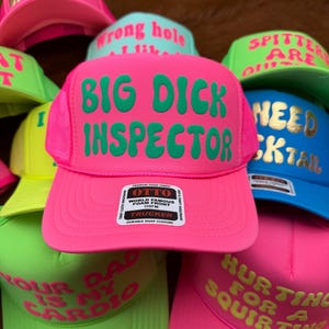 May include: A collection of trucker hats in various neon colors, including pink, green, and blue. Each hat features bold, humorous text in contrasting colors. The hats have a mesh back and a foam front, with a snap closure.